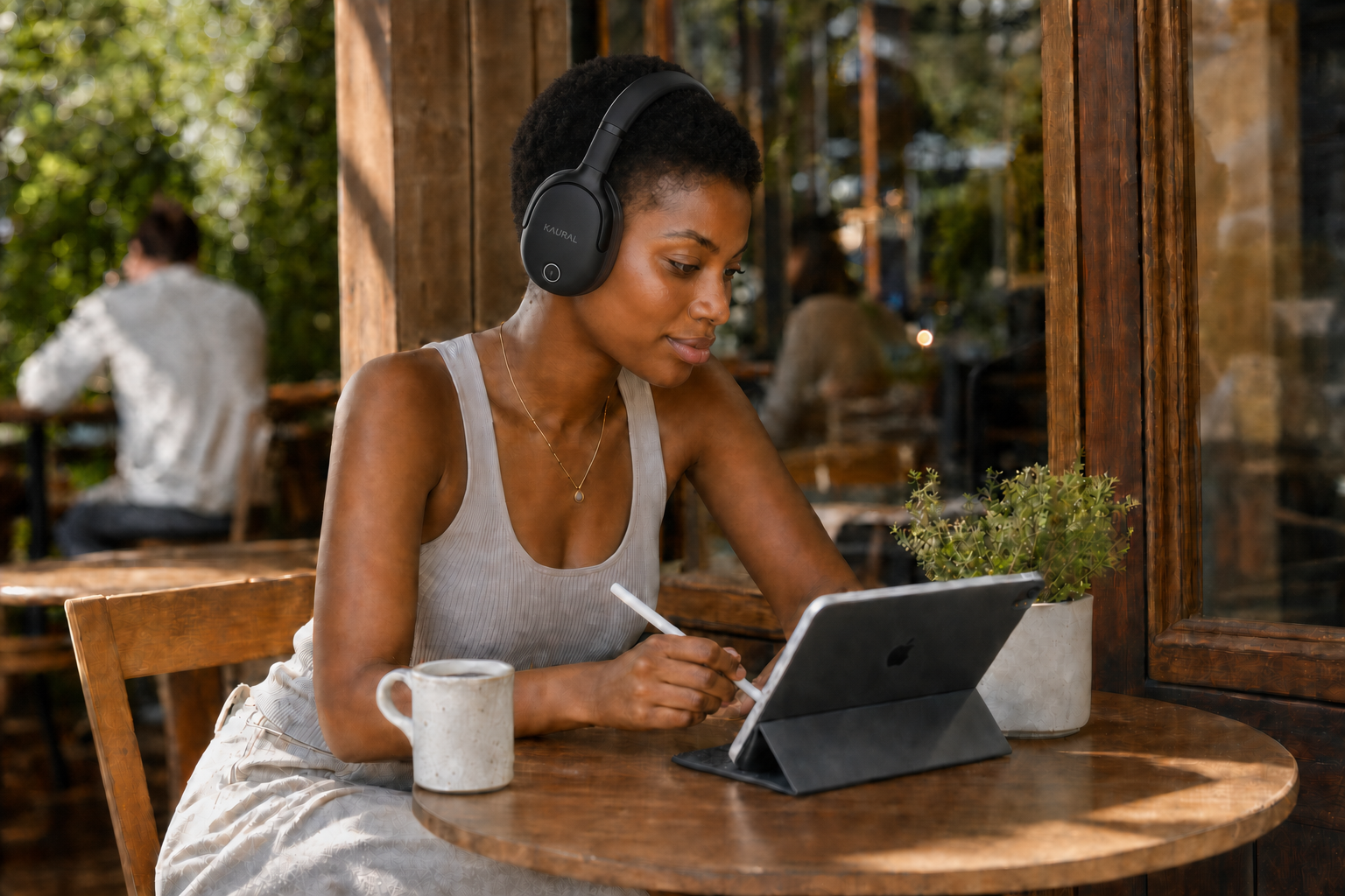 A woman wearing black over-ear headphones sits at a wooden table in a cafe, using a tablet with a stylus. A white mug is on the table next to her.